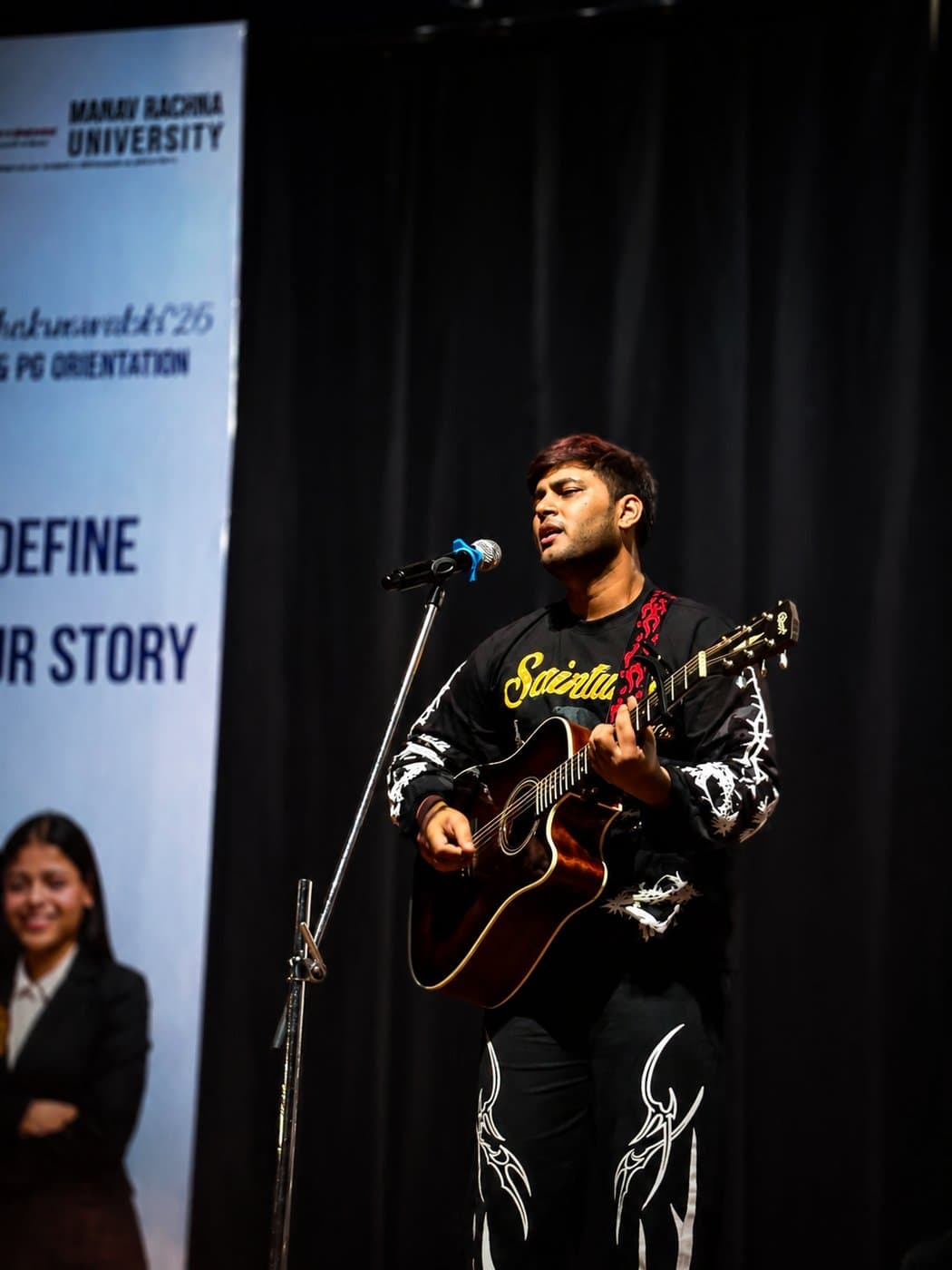 Hariank singing and playing acoustic guitar on stage at a university event.